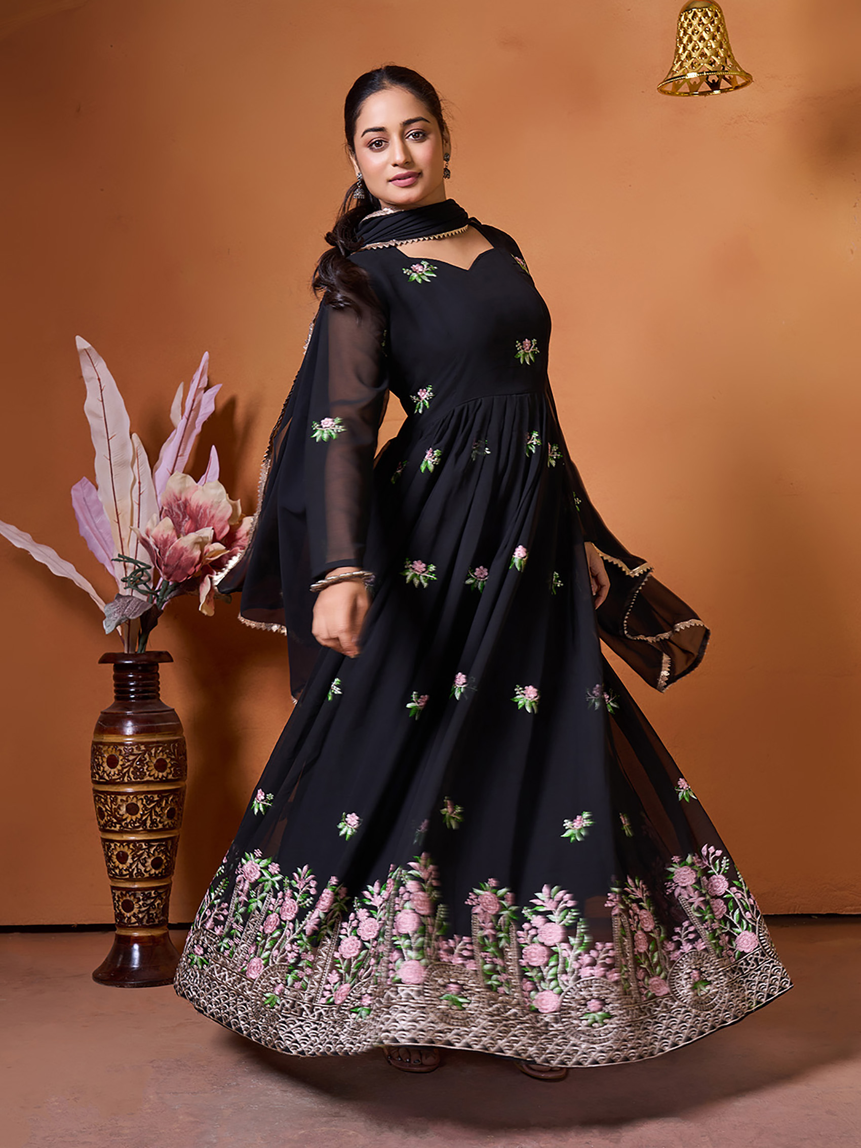 A model posed in a warm-toned studio setting wearing a black embroidered gown with traditional jewelry.