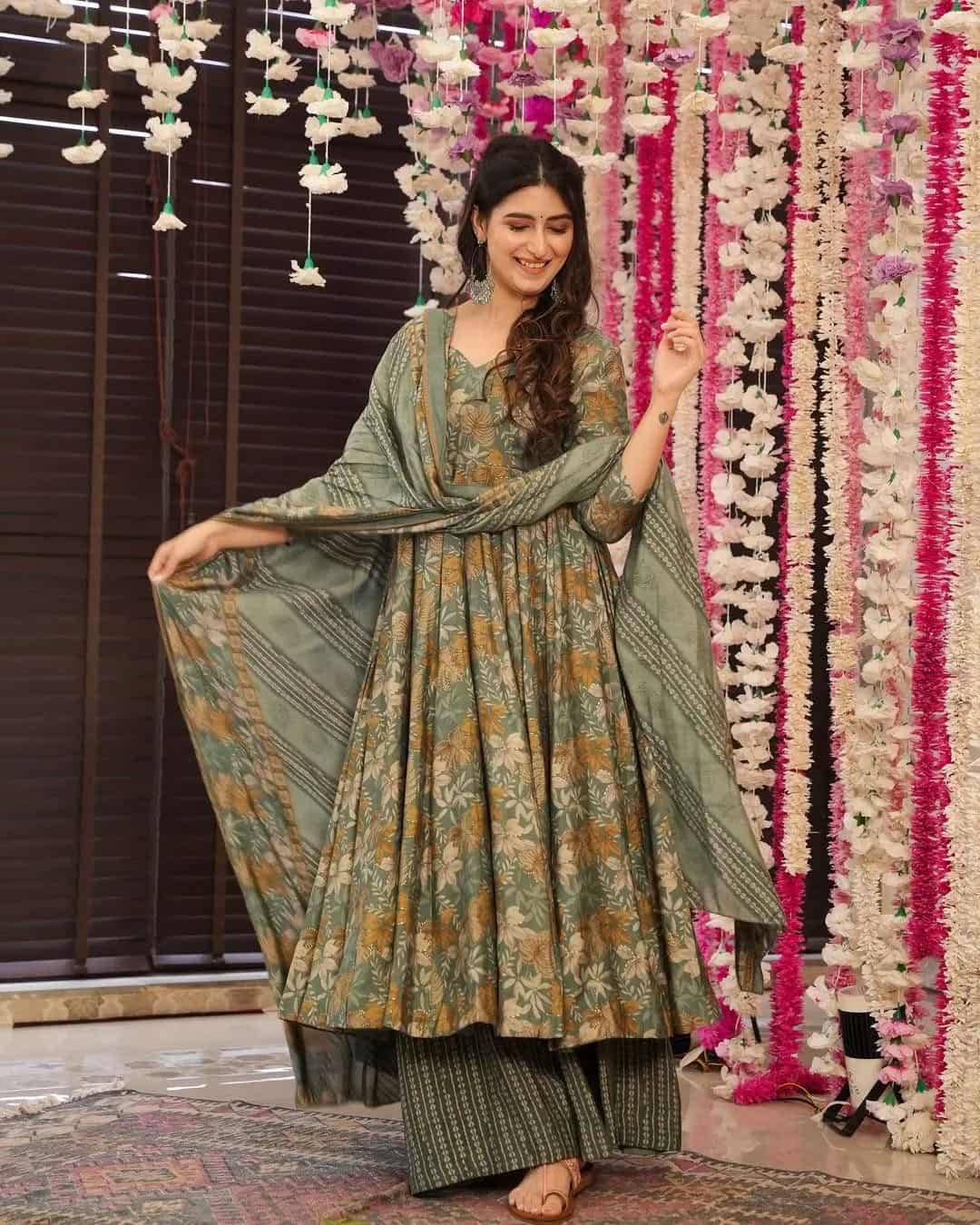 A woman posing in a flared olive green Anarkali featuring earth-toned floral prints and a matching dupatta with striped palazzo pants.